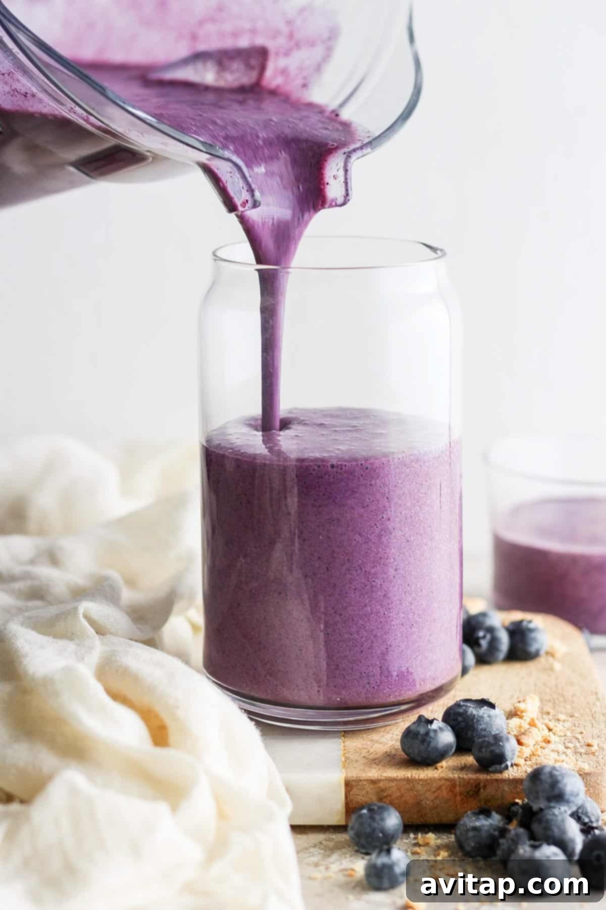 Freshly blended blueberry protein smoothie being poured into a clear glass jar, with scattered fresh blueberries on a rustic wooden board.