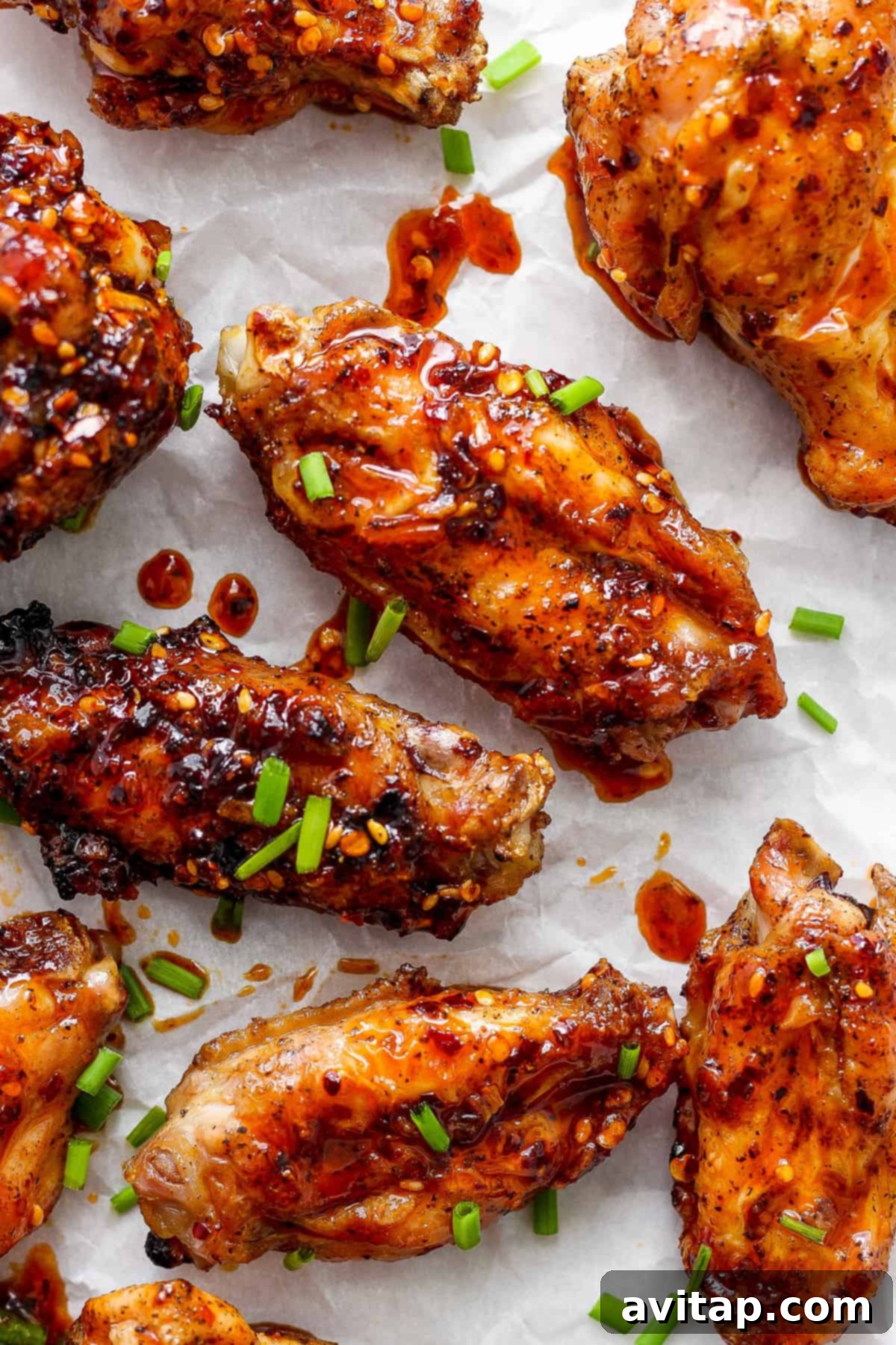 Close-up of glossy honey chili crisp grilled chicken wings, generously sprinkled with white sesame seeds and fresh chopped green onions, resting on parchment paper.