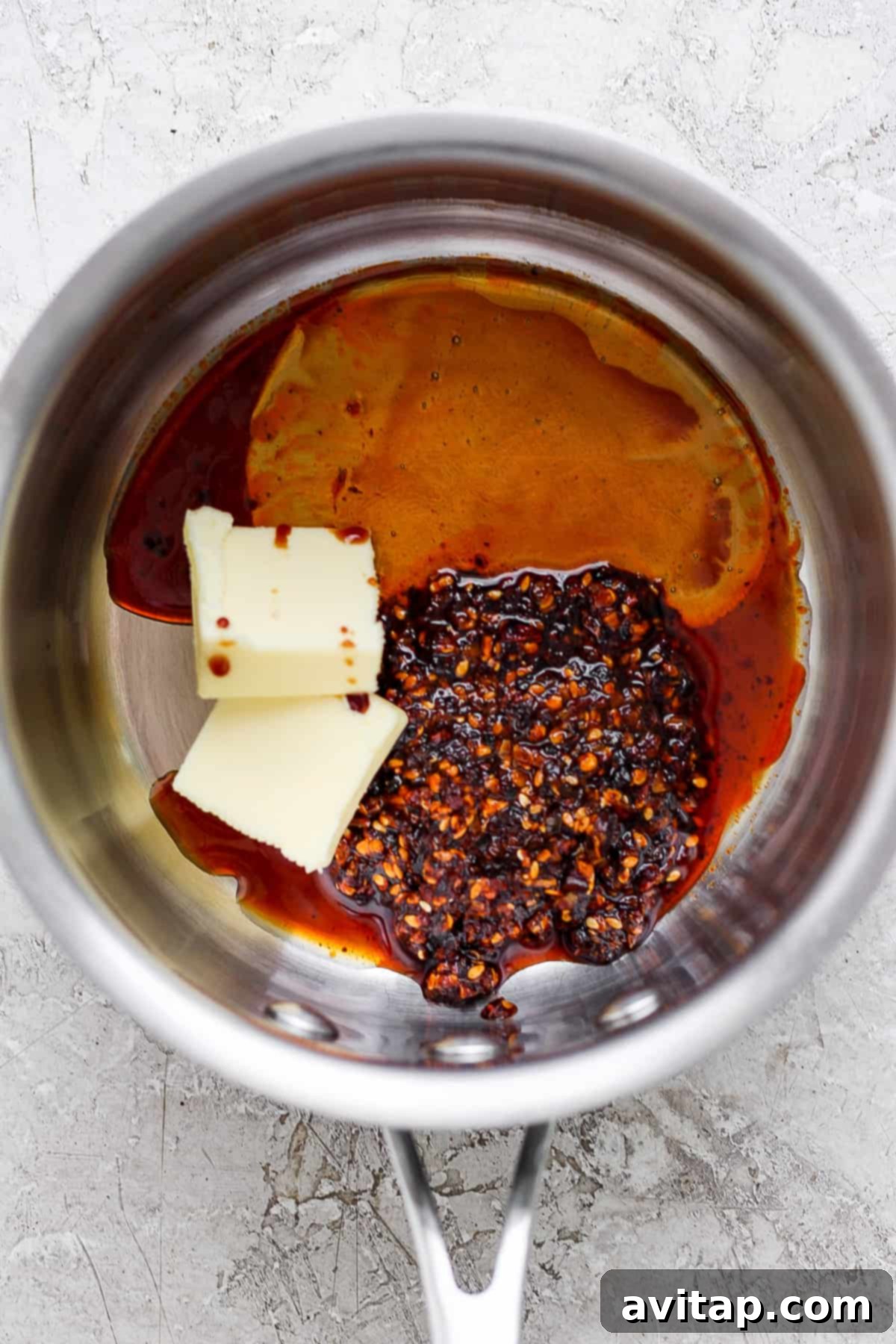 Close-up of various ingredients for honey chili crisp sauce simmering in a small stainless steel saucepan on a stove, including melted butter, vibrant chili flakes, and a dark, glossy liquid, hinting at the rich flavors being developed.