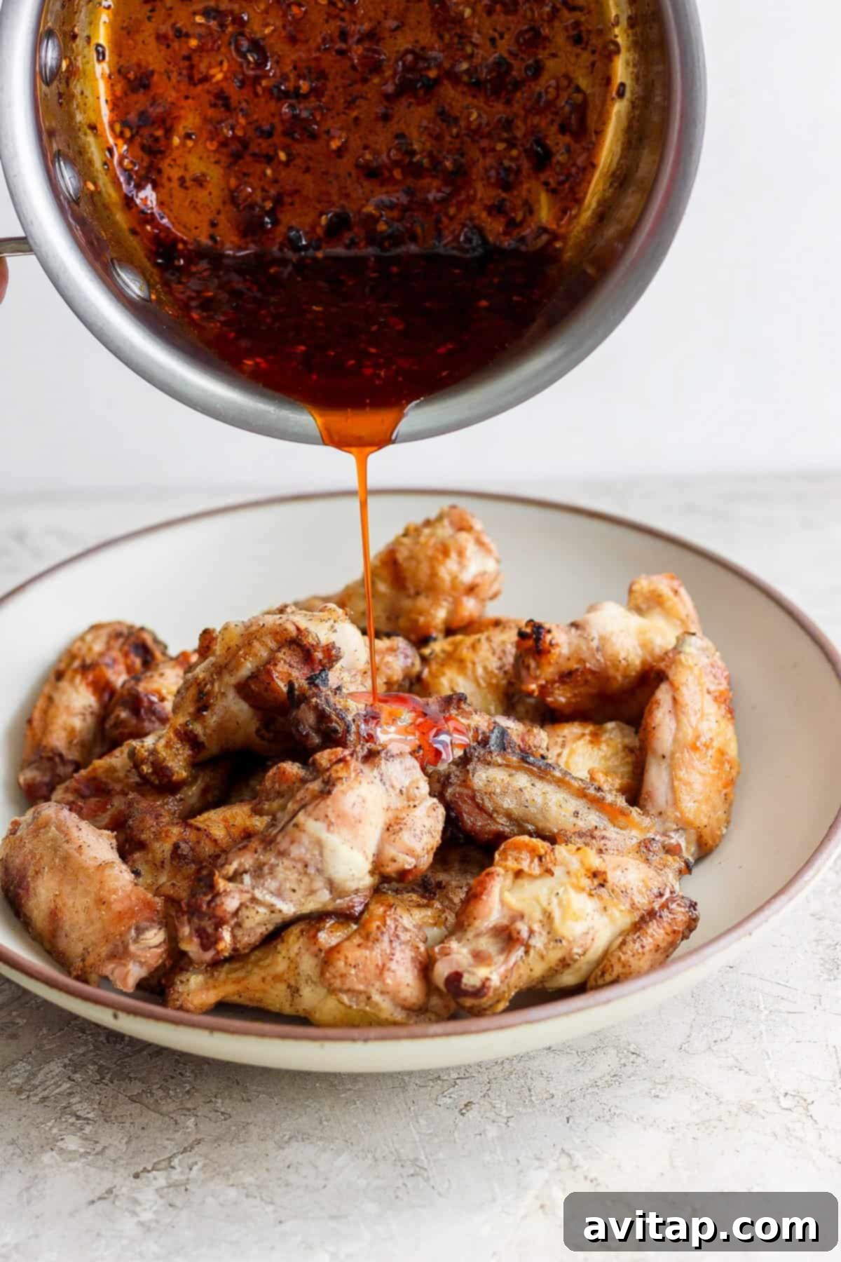 A generous cascade of rich, glossy honey chili crisp sauce being poured over a mound of freshly grilled chicken wings arranged on a serving plate.