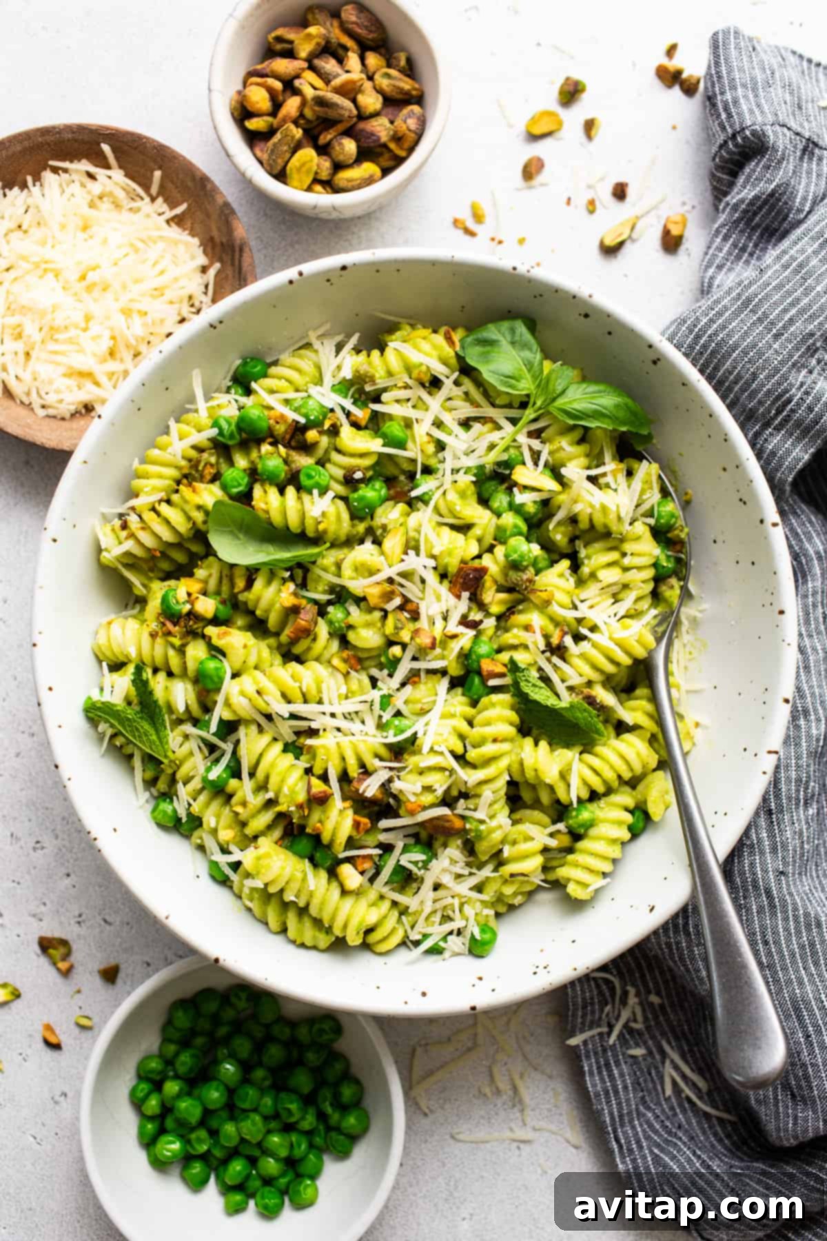 Fresh Sweet Pea Pesto Pasta 2 A vibrant bowl of Sweet Pea Pesto Pasta garnished with grated cheese and chopped pistachios, with fresh peas and basil leaves artfully arranged on the side.