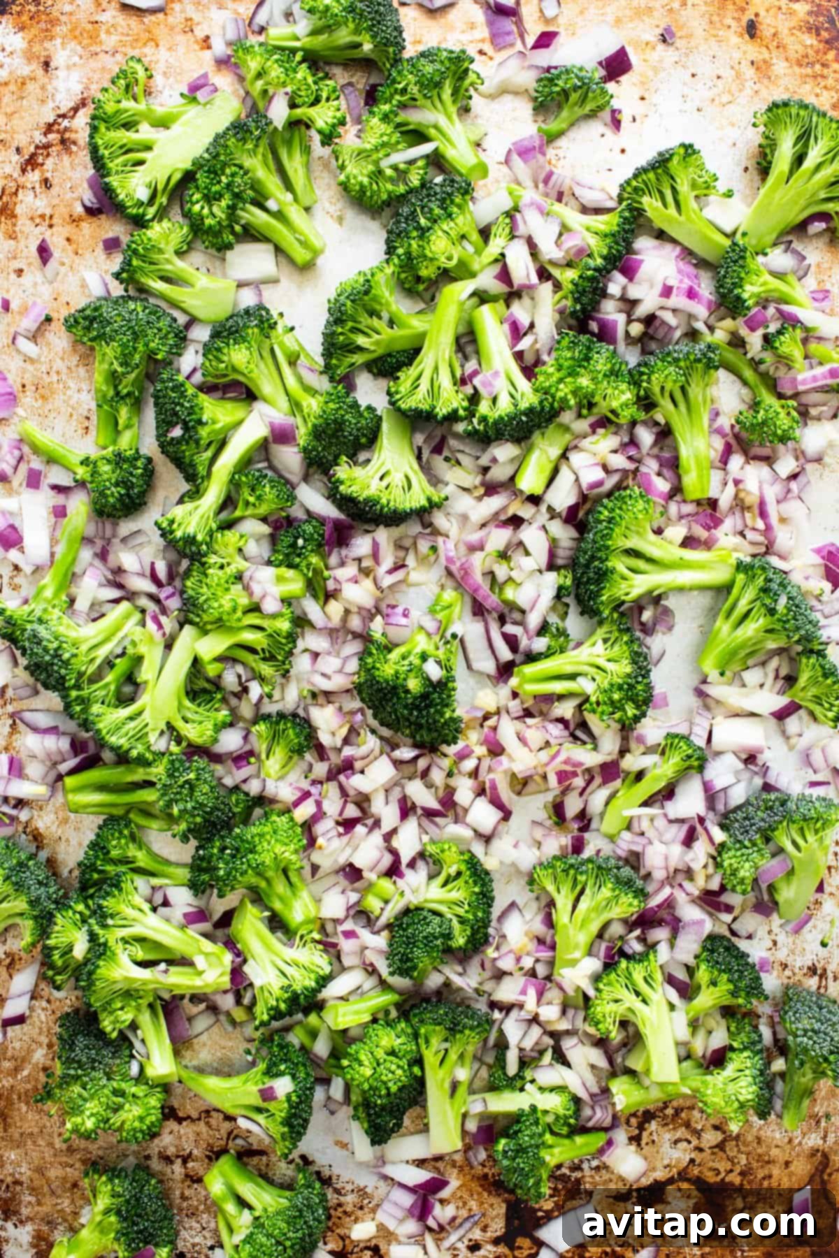 Freshly chopped broccoli florets and thinly sliced red onions are spread evenly on a parchment-lined baking sheet, glistening with olive oil and ready for roasting.