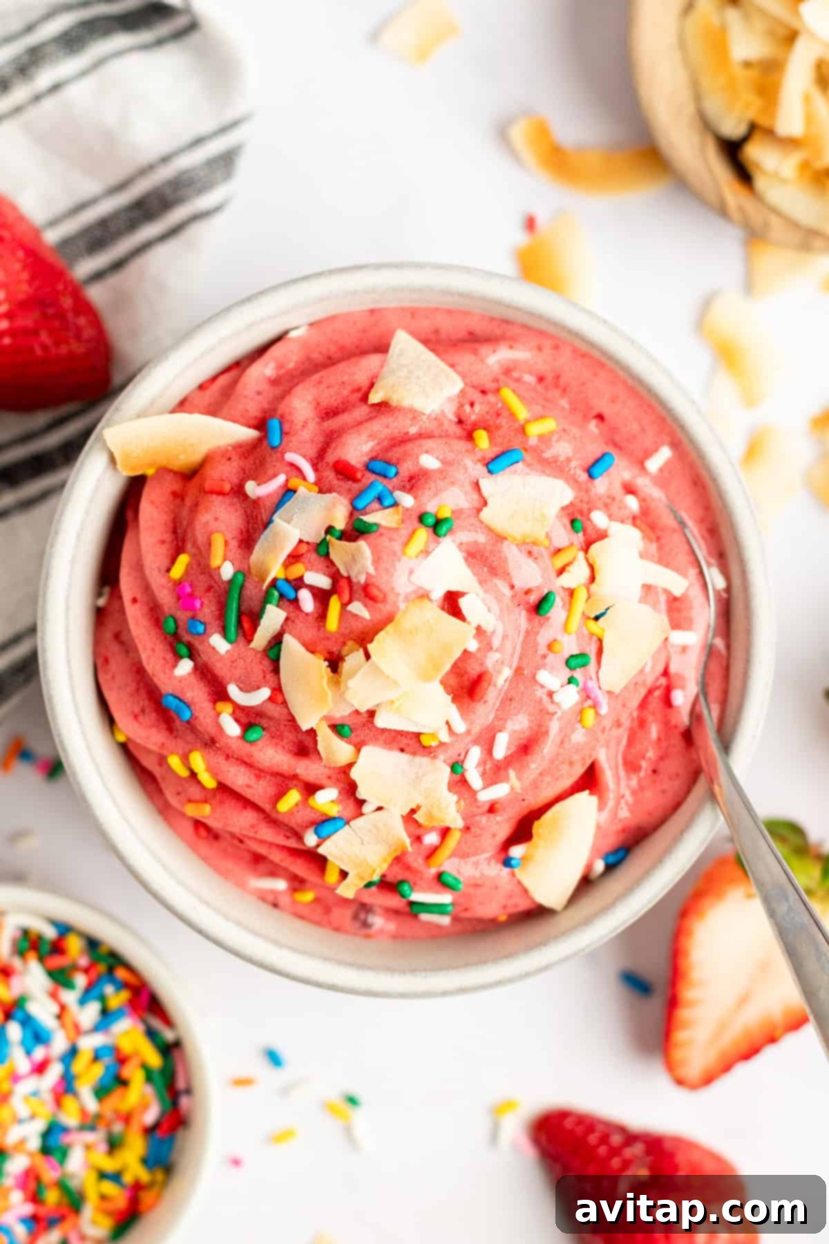 Strawberry Dole Whip 2 A vibrant pink smoothie bowl topped with coconut flakes and rainbow sprinkles, accompanied by fresh strawberries and a spoon, showcasing a delicious Strawberry Dole Whip.
