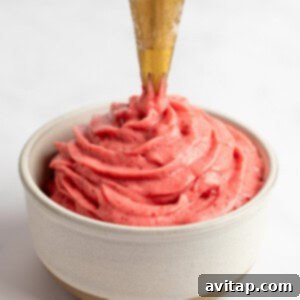 A piping bag dispensing creamy strawberry frosting into a small beige bowl against a white background, showing the soft-serve consistency being achieved.