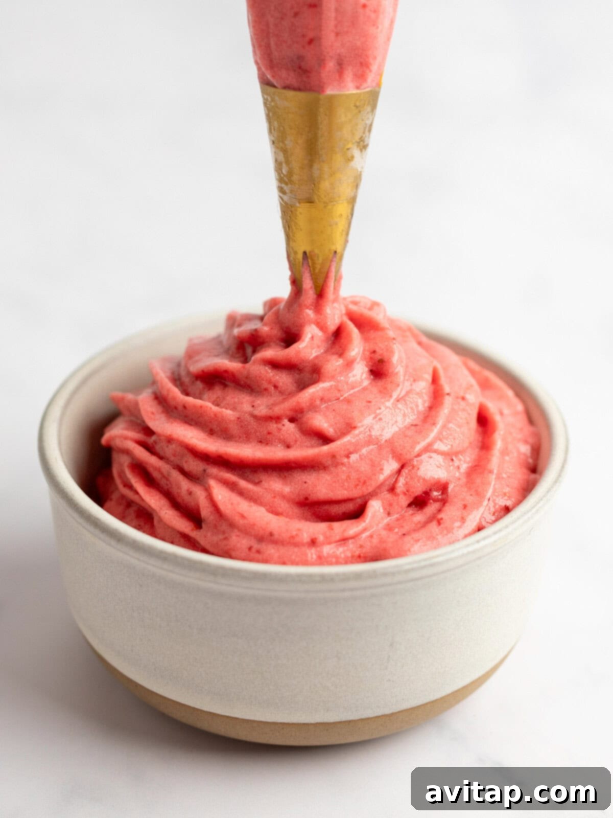 Strawberry Dole Whip 7 A piping bag dispensing creamy strawberry frosting into a small beige bowl against a white background, demonstrating the ideal consistency for serving.