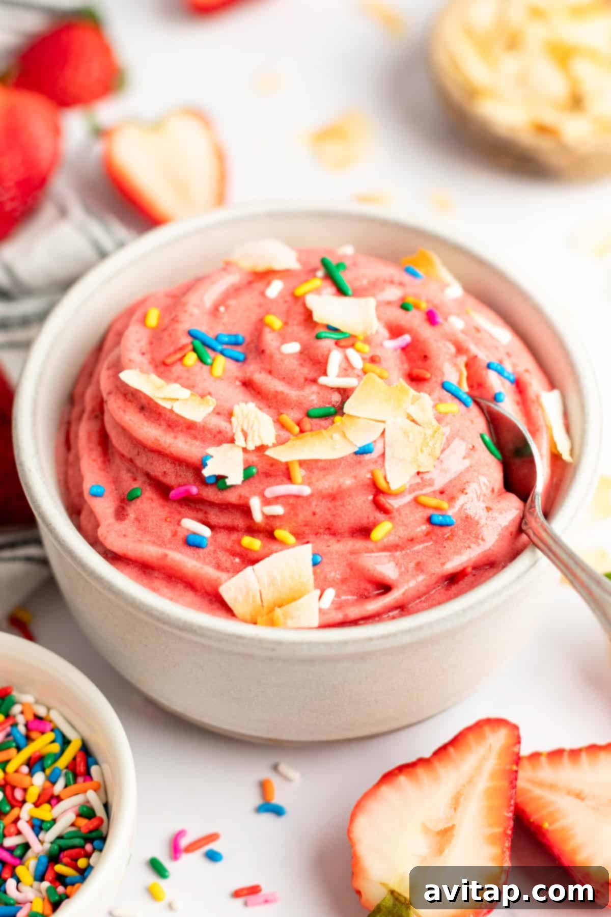 Strawberry Dole Whip 8 A bowl of pink strawberry banana smoothie bowl topped with sprinkles and coconut flakes, surrounded by fresh strawberries and sprinkles, showcasing a delightful dessert.