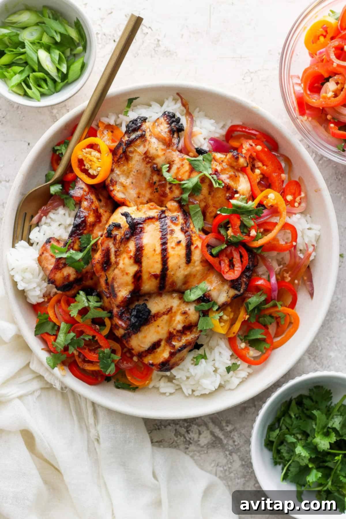 Tropical Grilled Coconut Chicken 2 Grilled chicken thighs on a bed of white rice, garnished with sliced bell peppers, onions, and herbs in a bowl, with chopsticks on the side.