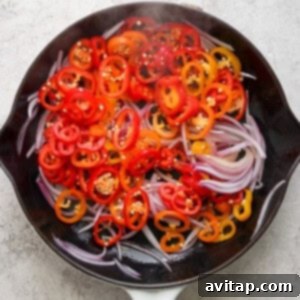 Sliced red and yellow bell peppers with red onions in a white le creuset skillet on a textured surface.