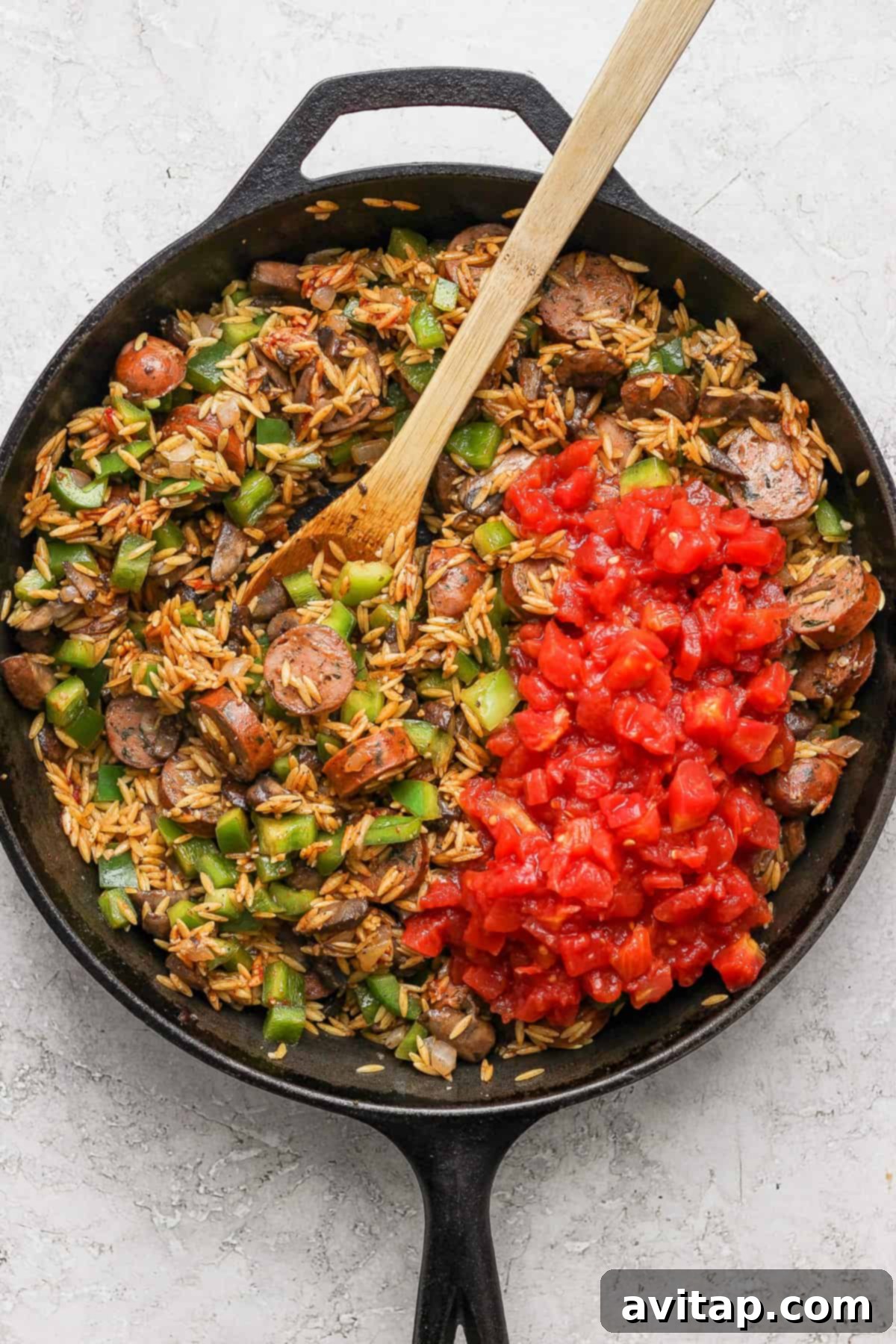 A skillet full of orzo pasta and chicken sausage with a wooden spoon.