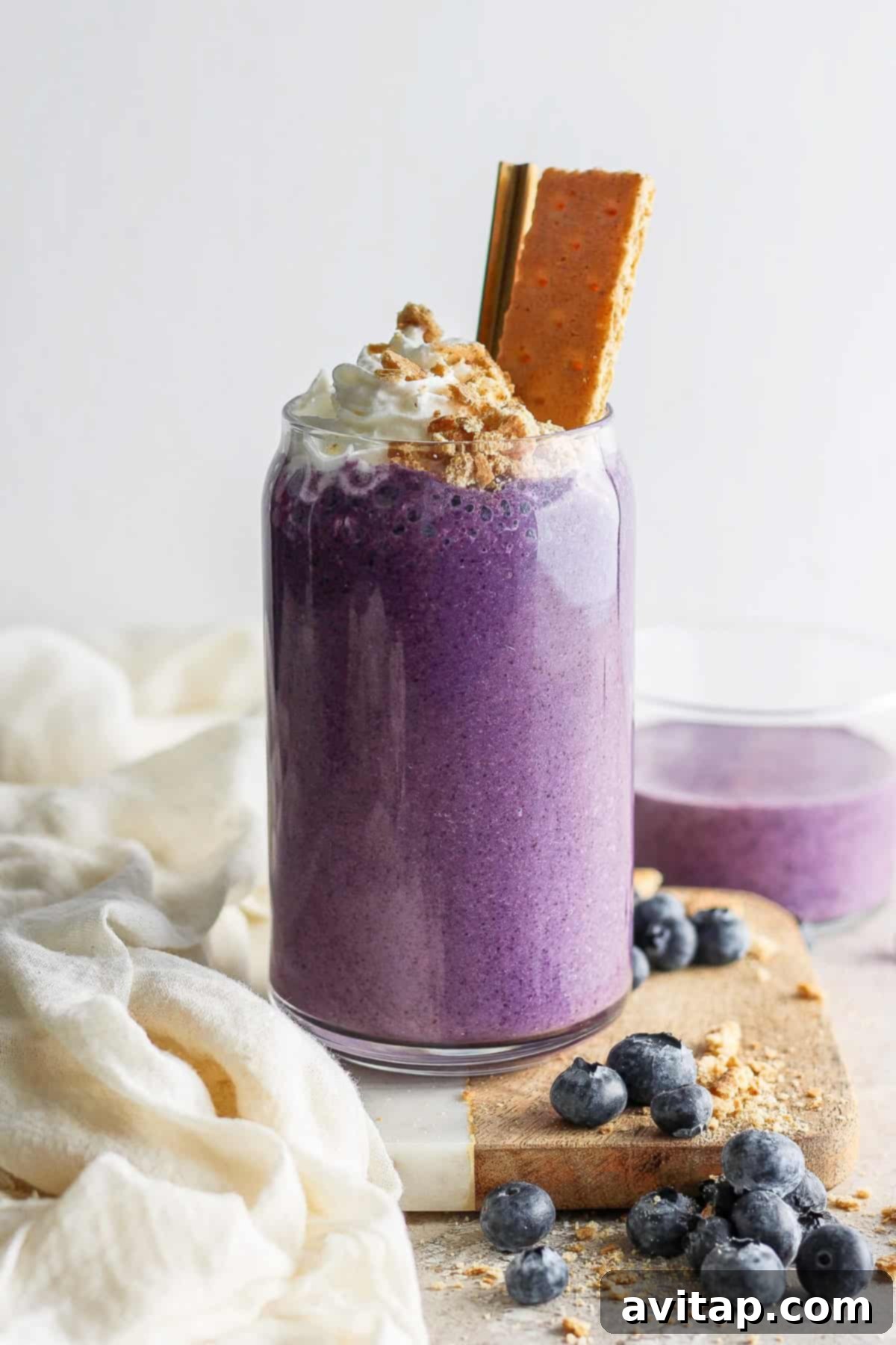 A vibrant purple smoothie topped with whipped cream and a cookie, garnished with blueberries on a light, textured background.