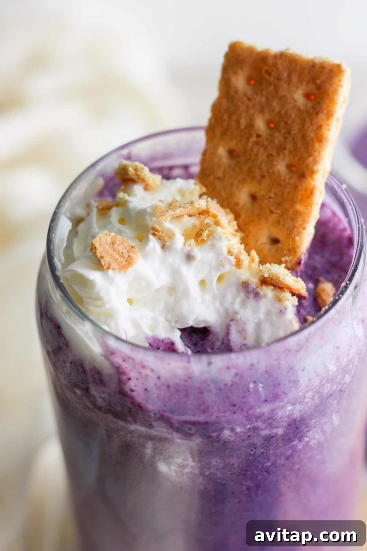 A close-up of a purple milkshake topped with whipped cream, crumbled cookies, and a cookie sticking out, served in a clear glass.