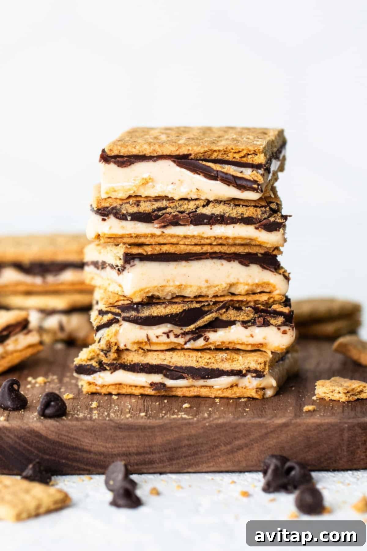 A stack of s'mores bars with layers of graham crackers, melted marshmallow, and chocolate on a wooden board, with scattered chocolate chips around.