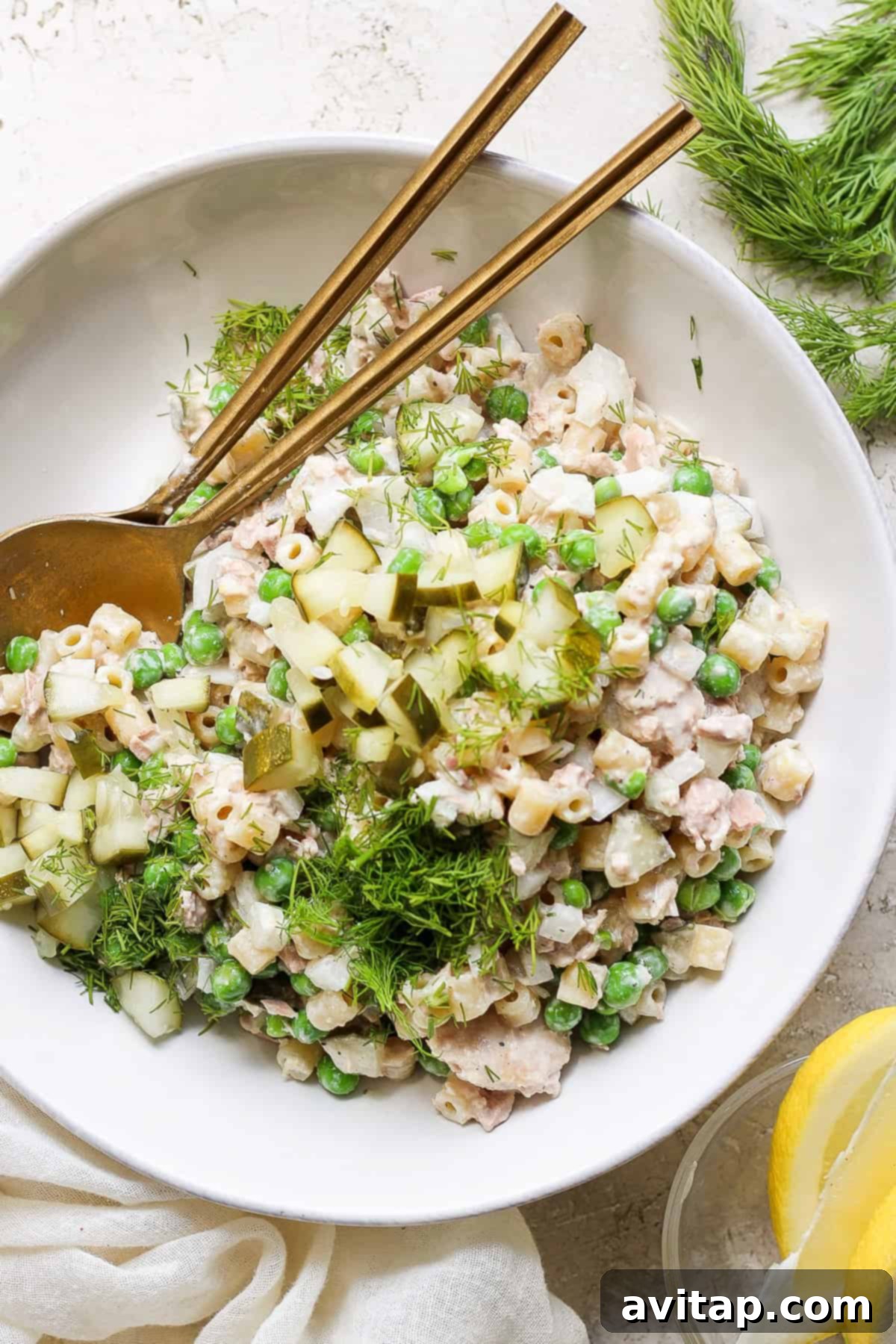 A bowl of tuna salad with peas, chopped cucumber, and dill, served with a pair of chopsticks and fresh lemon slices on a neutral-toned background.