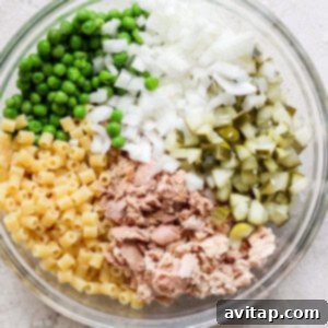 A bowl containing separate sections of diced onions, green peas, chopped pickles, macaroni pasta, and canned tuna, arranged on a light, textured surface.