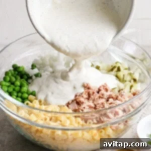 A person pours creamy sauce into a bowl containing macaroni, tuna, chopped onions, and peas for a salad.