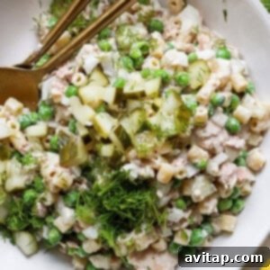 A bowl of tuna salad with peas, chopped cucumbers, and dill, served with a pair of gold chopsticks on a white plate.