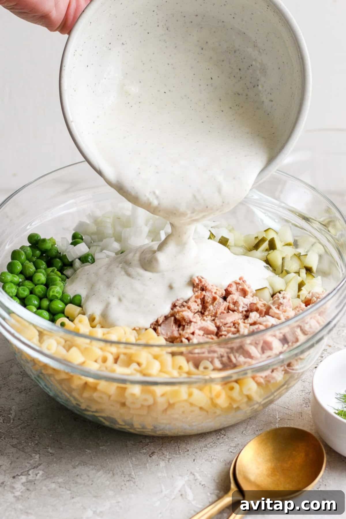 A person pours creamy sauce into a bowl containing macaroni, tuna, chopped onions, and peas for a salad.
