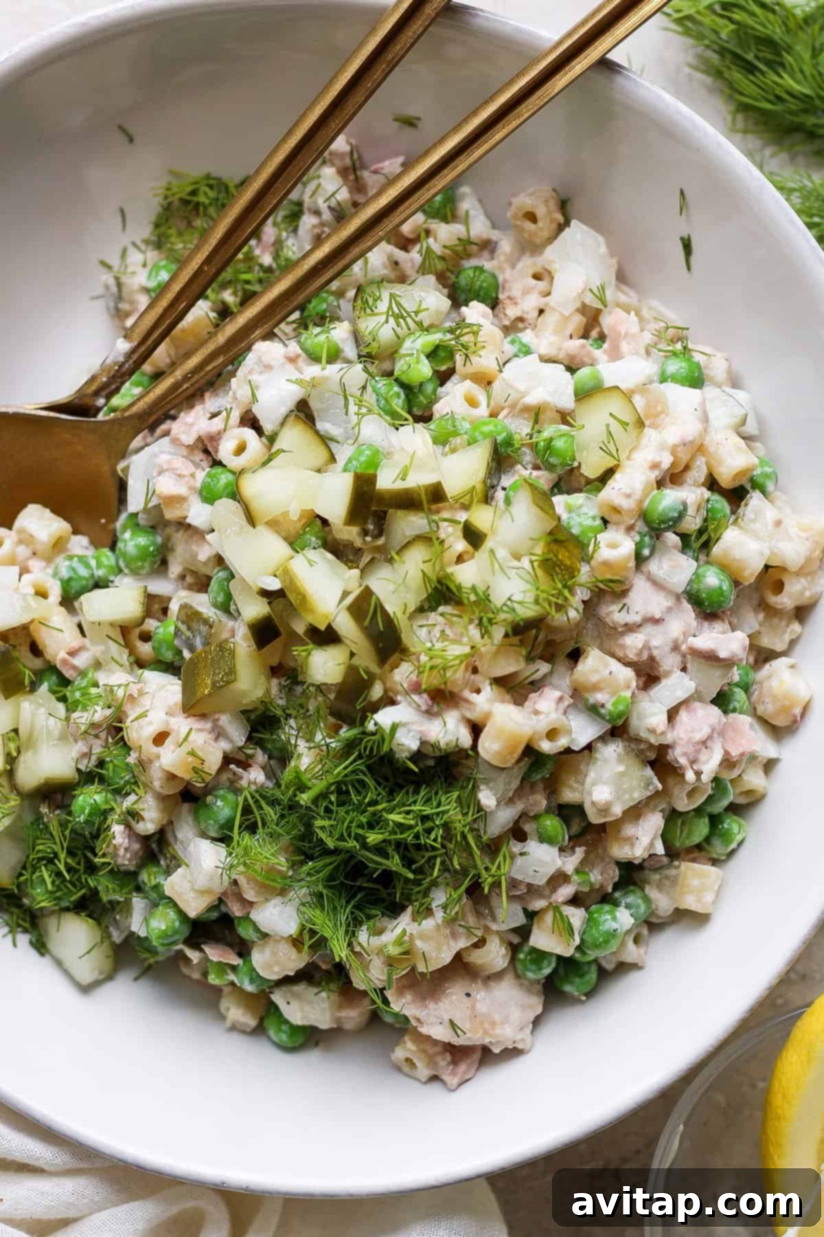 A bowl of tuna salad with peas, chopped cucumbers, and dill, served with a pair of gold chopsticks on a white plate.