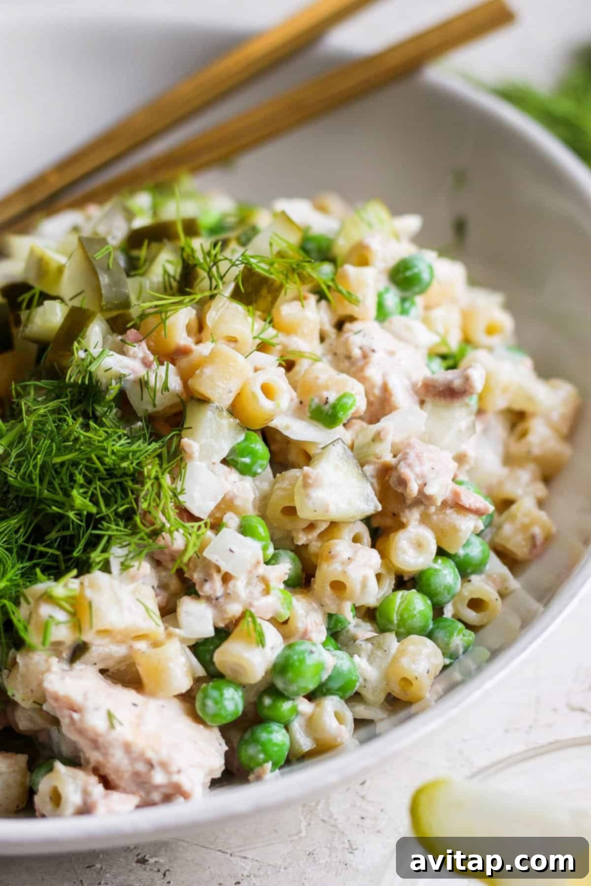 A bowl of tuna pasta salad with peas and dill, garnished with fresh herbs and served with chopsticks.