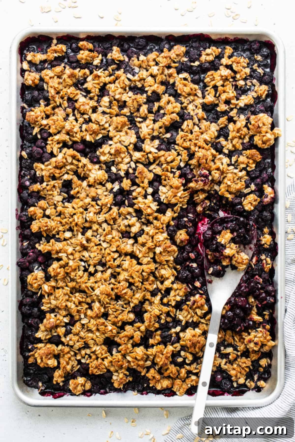 A baked blueberry oatmeal crumble in a rectangular pan with a serving spoon, showcasing its golden, crispy topping.