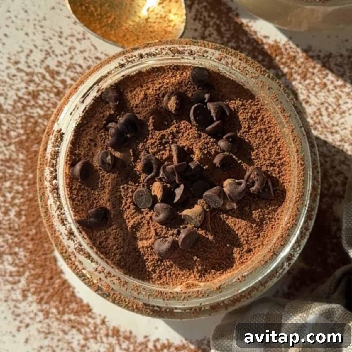 A jar filled with a powdered brown substance topped with chocolate chips hints at the delicious promise of blended overnight oats. A gold spoon is partially visible nearby, with more powder scattered on the surface, ready to be enjoyed.