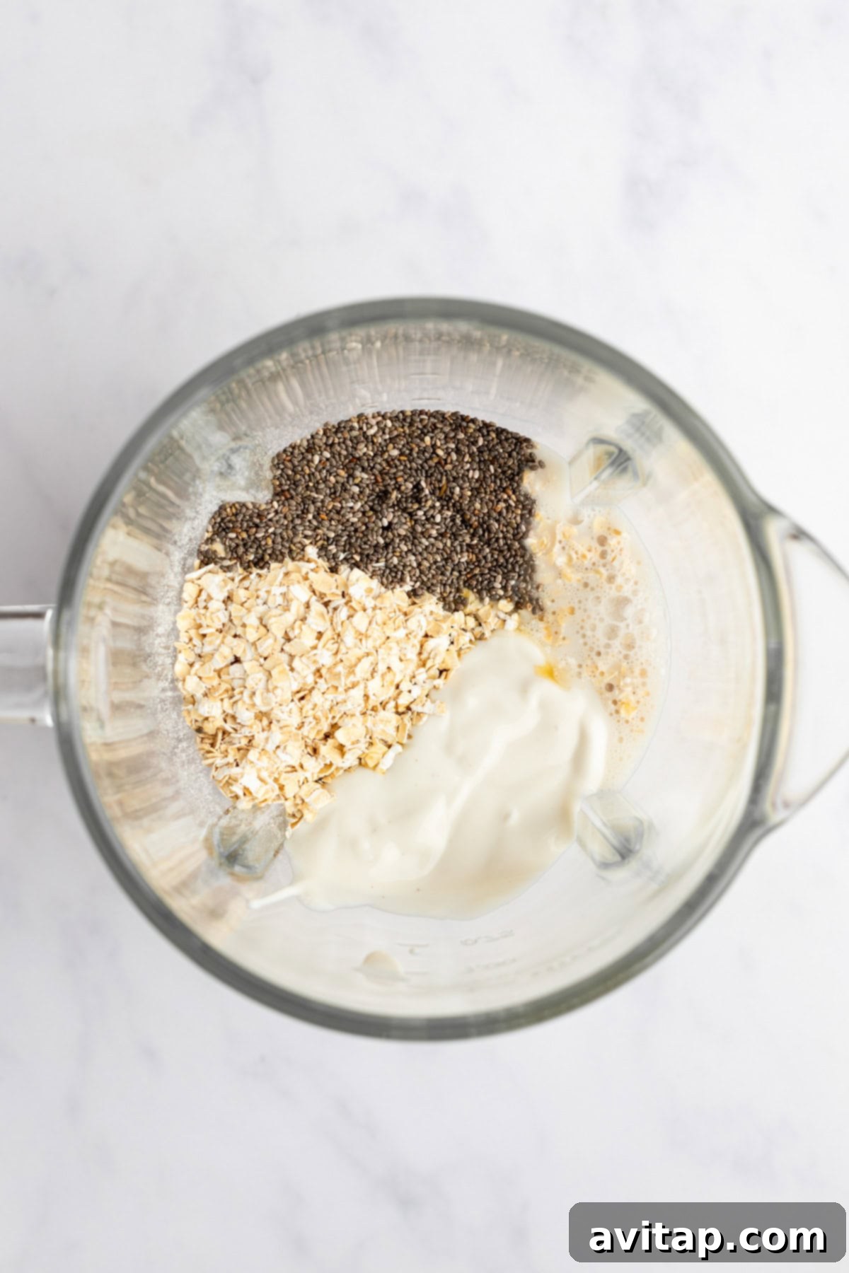 A blender containing chia seeds, oats, a white creamy substance, and liquid viewed from above on a light-colored surface, ready for blending.