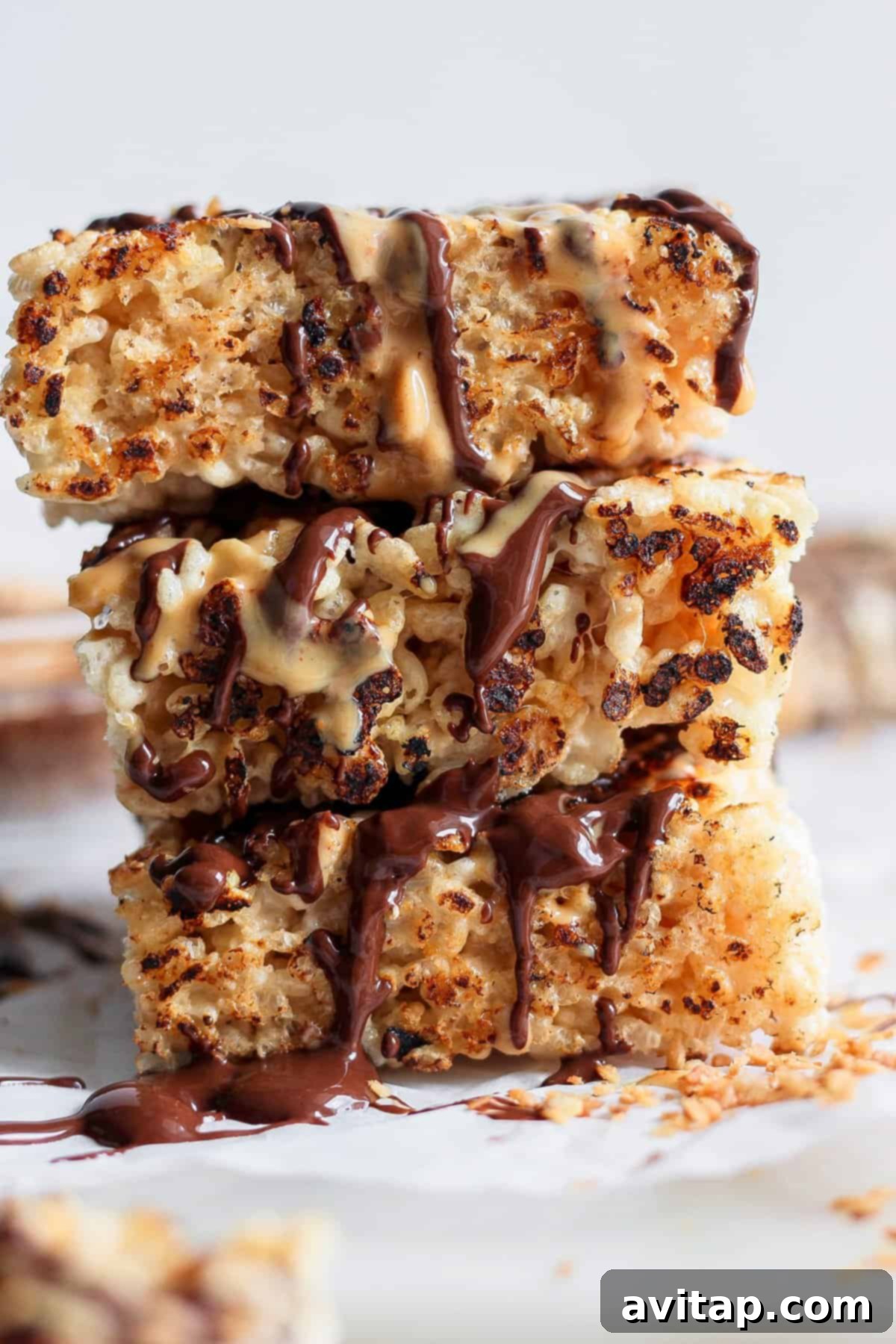 Stack of three toasted rice Krispie treats drizzled with chocolate and peanut butter on a white surface, garnished with chopped nuts.
