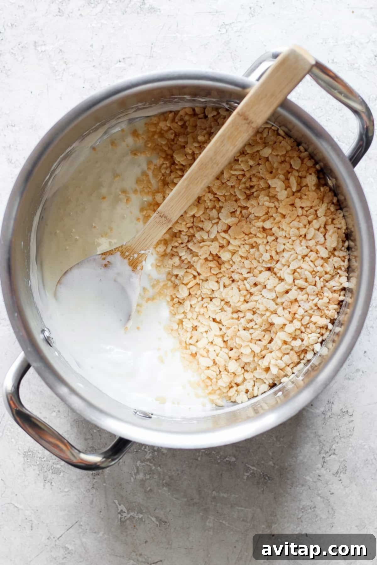 A pot filled with melted marshmallows and some toasted Rice Krispie treats, with a wooden spoon resting inside, on a light-colored surface, demonstrating the preparation process.