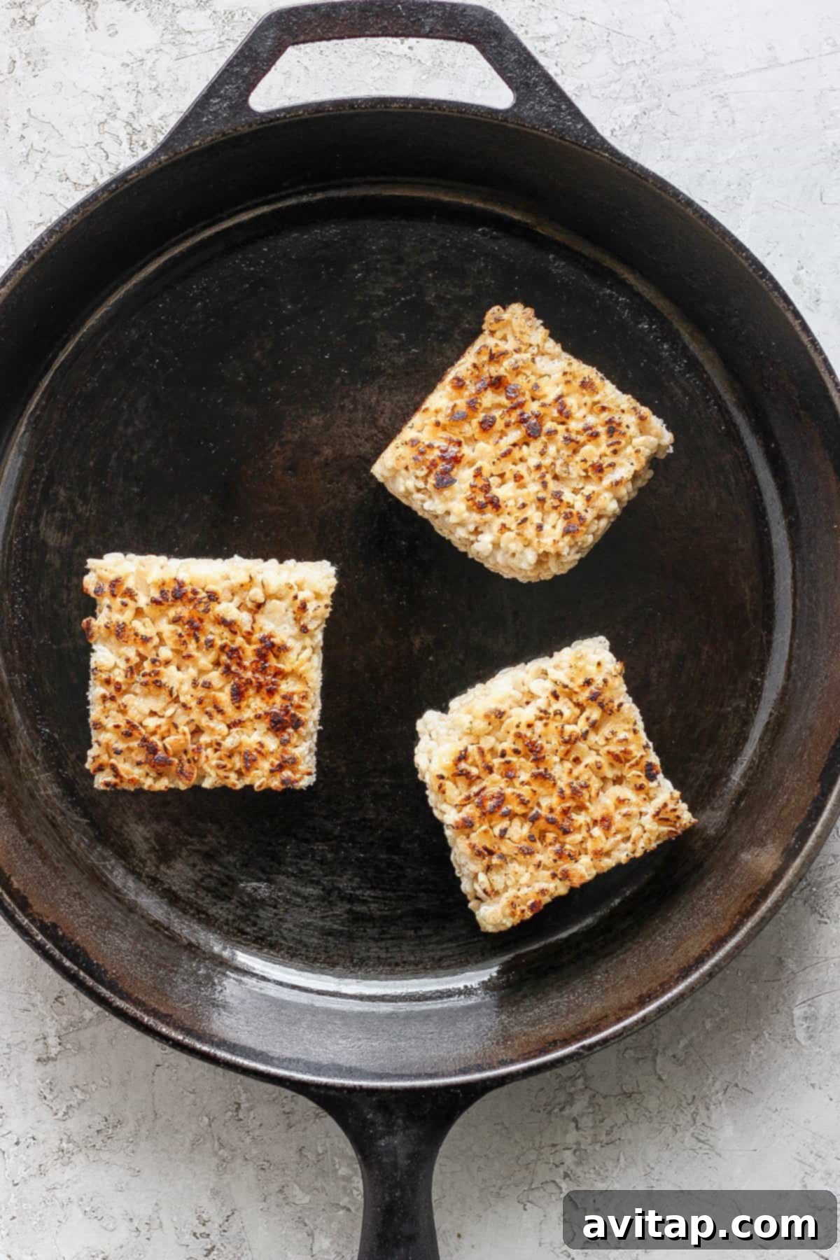 Three toasted Rice Krispie treats in a cast iron skillet on a light textured surface, highlighting their golden-brown finish.