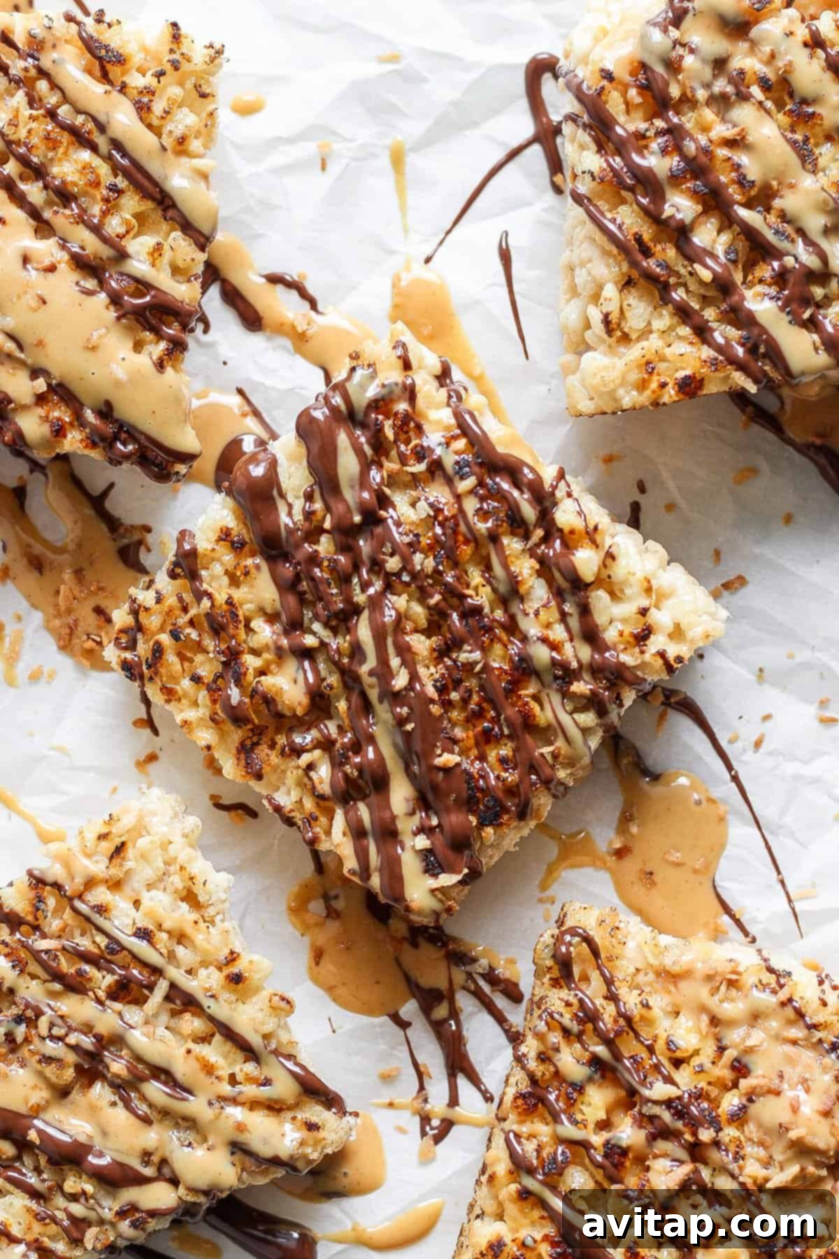 Close-up of five toasted Rice Krispie treats drizzled with chocolate and peanut butter, arranged on parchment paper and topped with crushed nuts.