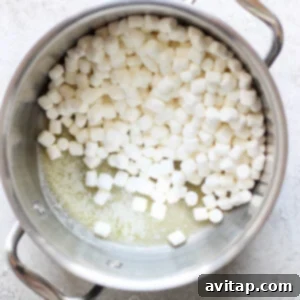 A metal pot containing partially melted mini marshmallows on a light-colored surface, ready to be transformed into delicious toasted Rice Krispie treats.