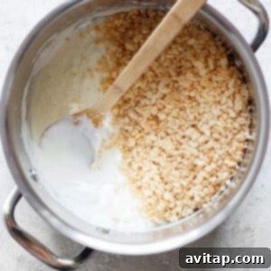 A pot filled with melted marshmallows and some toasted Rice Krispie treats, with a wooden spoon resting inside, on a light-colored surface, illustrating the mixing stage.
