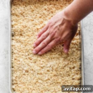A hand pressing down on a tray filled with warm, toasted Rice Krispie treats mixture, ensuring an even layer.