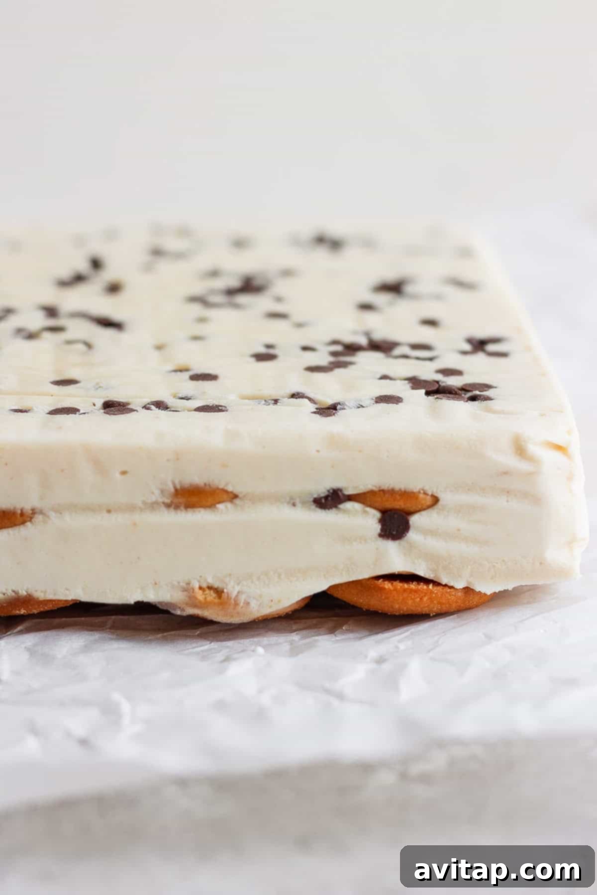 Peanut Butter Banana Icebox Delight 4 Close-up of a layered peanut butter banana ice box cake with a creamy white filling, chocolate chips on top, and a base of cookies, set on crinkled parchment paper.