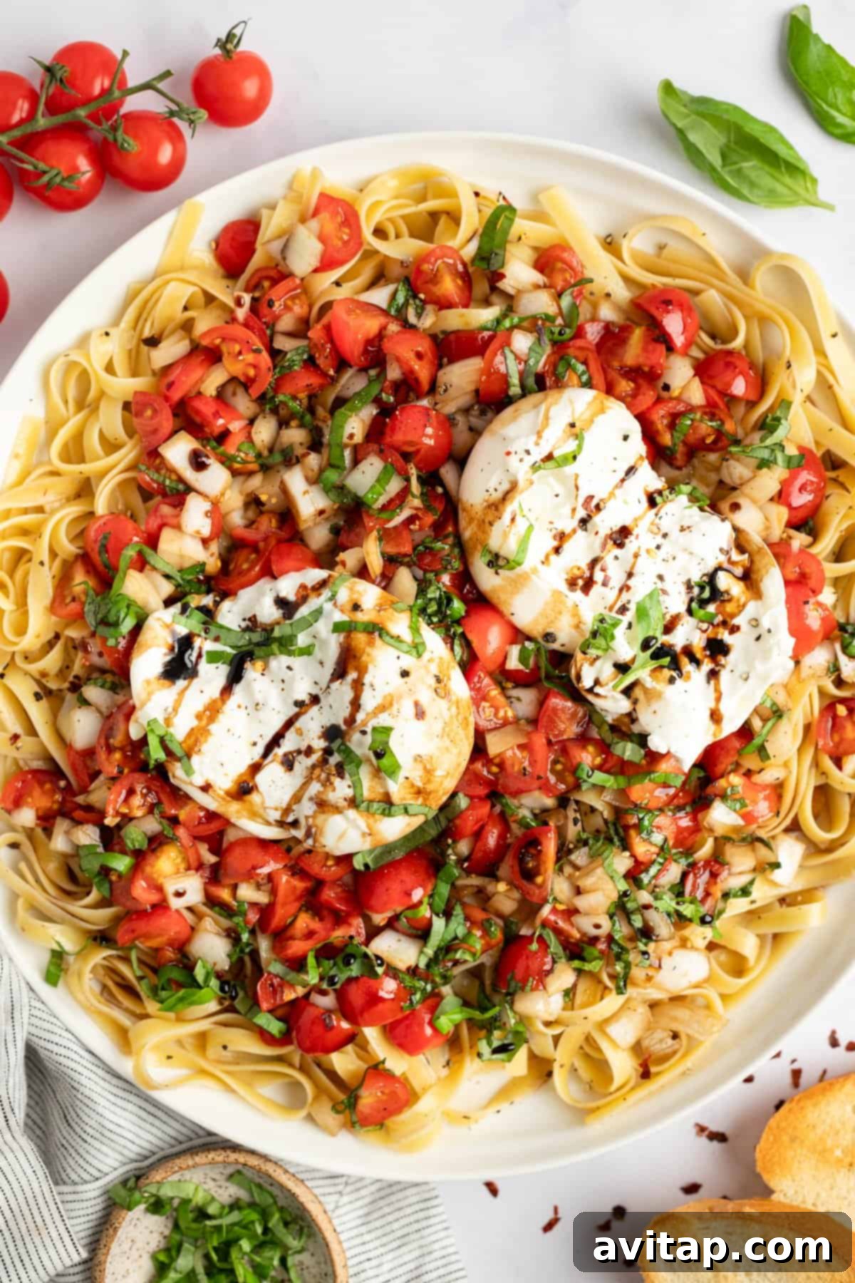 A plate of fettuccine pasta topped with fresh chopped tomatoes, basil, and a drizzle of balsamic glaze, garnished with a generous dollop of burrata cheese and black pepper.