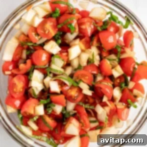 A glass bowl filled with a fresh salad of chopped tomatoes, cucumbers, and herbs on a white surface.