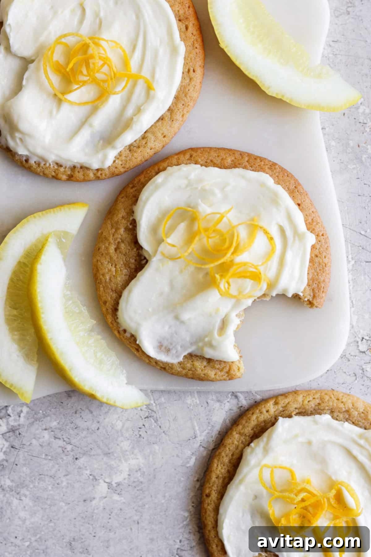 Sunshine Lemon Cream Cheese Cookies 2 Three lemon cookies with cream cheese frosting, topped with fresh zest, sit invitingly on a cutting board. One is partially eaten, revealing its soft interior. Beside them, lemon wedges are artfully arranged on the textured surface, enhancing the citrusy allure of the scene.