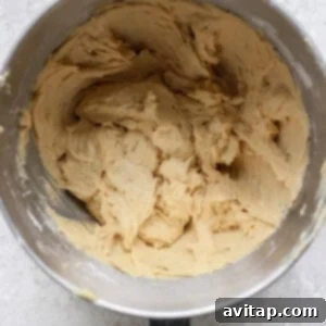 A mixing bowl filled with creamy, beige lemon cookie dough on a textured, light gray surface evokes the promise of sweet treats topped with luscious cream cheese frosting.