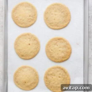 Six round lemon cookies with cream cheese frosting rest on a parchment-lined baking sheet.