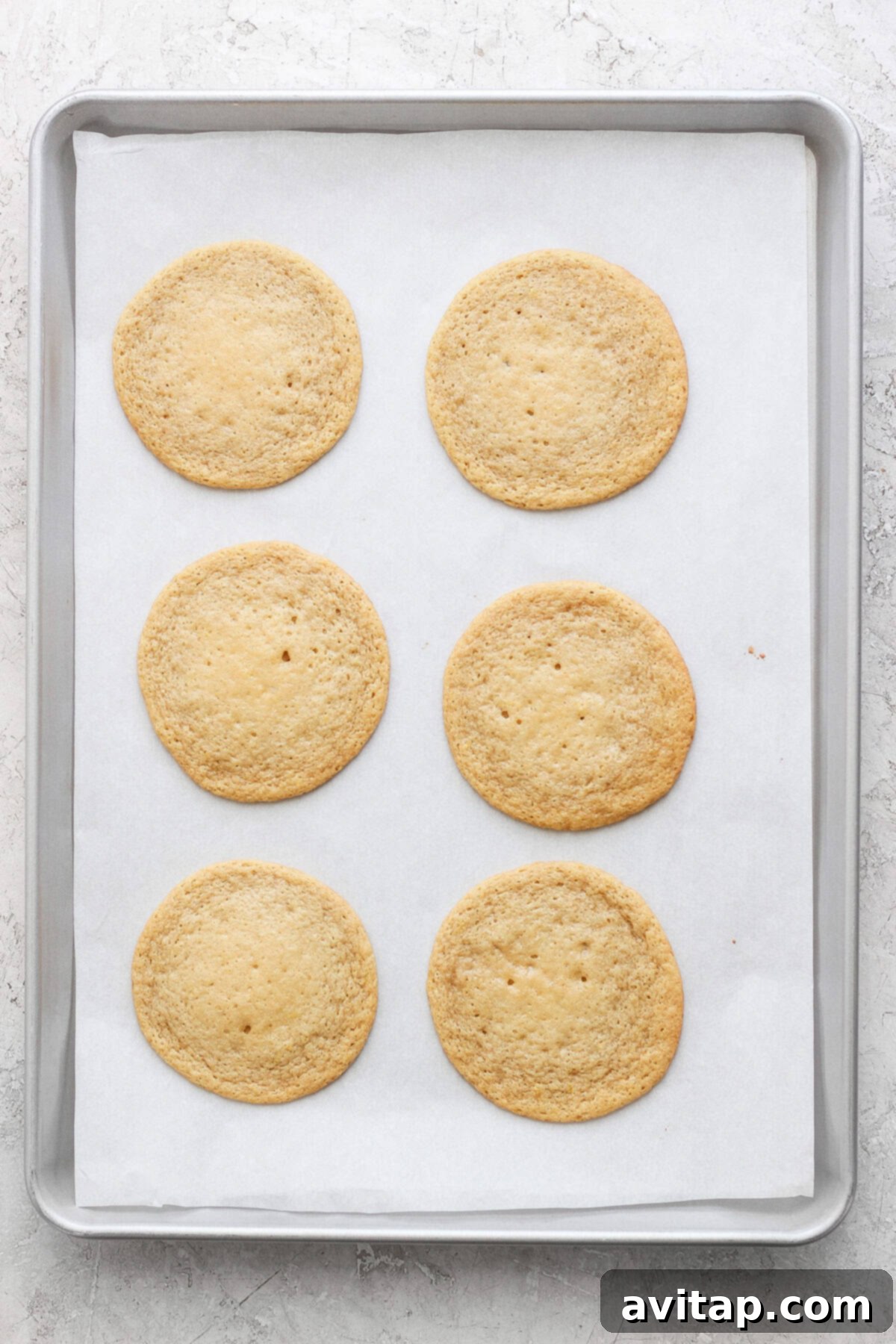 Sunshine Lemon Cream Cheese Cookies 4 Six round lemon cookies with cream cheese frosting rest on a parchment-lined baking sheet.