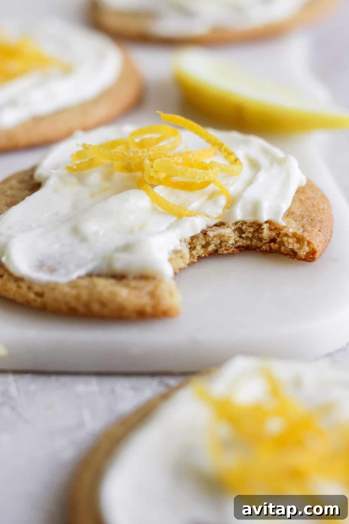 Sunshine Lemon Cream Cheese Cookies 6 Close-up of a lemon cookie with cream cheese frosting and lemon zest on top. A bite is taken out of the cookie, revealing its soft, chewy interior. A lemon wedge is visible in the background, emphasizing the fresh citrus flavor.