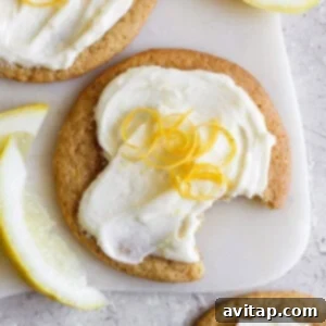 A lemon cookie with cream cheese frosting, topped with lemon zest, has a bite taken out of it. Slices of lemon are nearby on a light surface, highlighting the fresh ingredients.