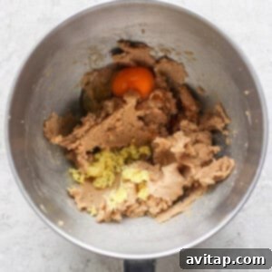 A mixing bowl brimming with cookie dough, a cracked egg, grated ginger, and hints of lemon zest prepares the stage for delightful lemon cookies with cream cheese frosting on a light textured surface.