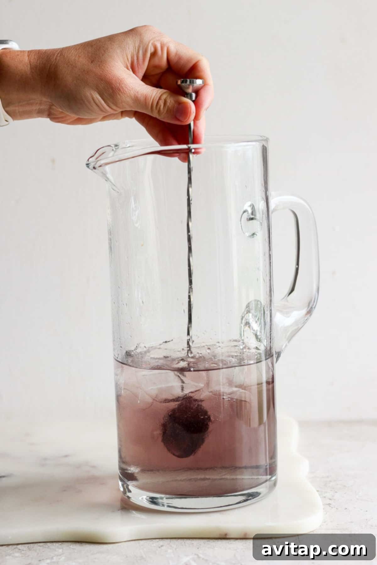 A hand stirring a purple liquid in a clear glass pitcher with ice, mixing the cocktail ingredients for a magical drink.