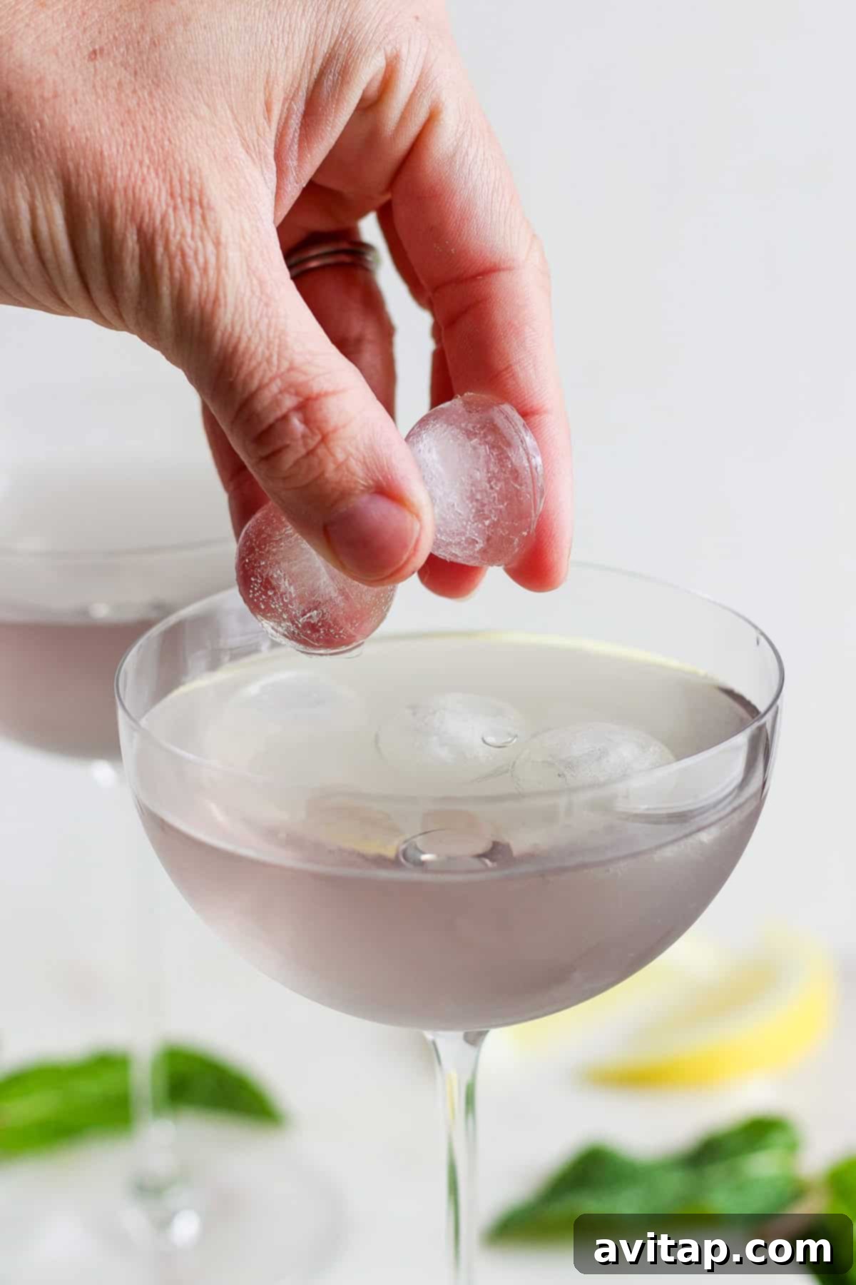 A hand gently placing fresh ice cubes into a cocktail glass filled with a clear, vibrant liquid, preparing for the magical color change.