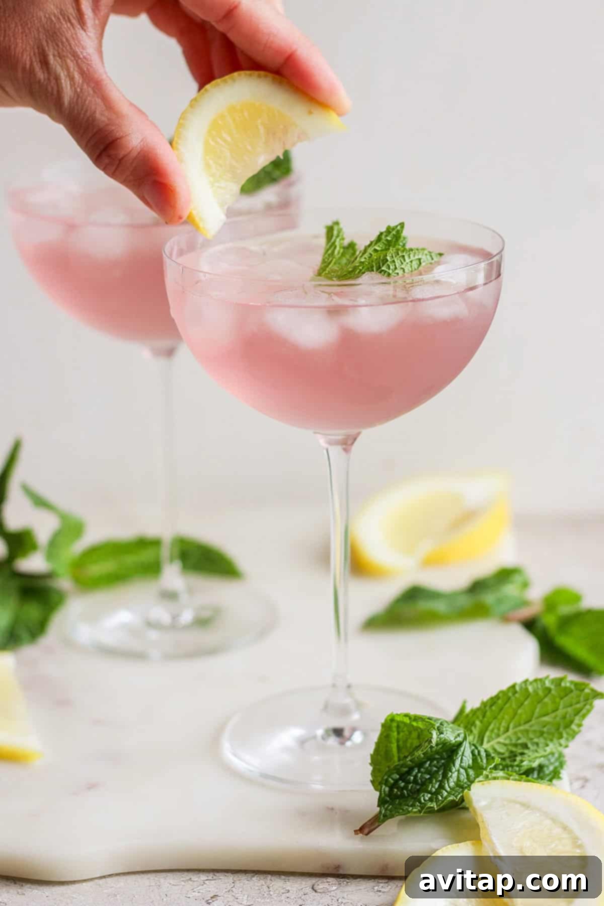 A hand placing a lemon wedge on the rim of a vibrant pink mocktail garnished with fresh mint leaves, showcasing the drink's transformation.