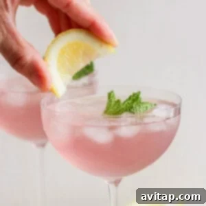 A hand placing a lemon slice on the rim of a vibrant pink cocktail garnished with fresh mint in a stemmed glass.