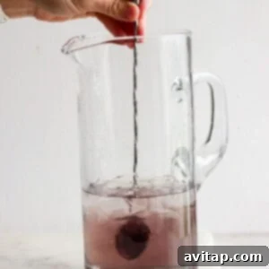 A hand stirring a vibrant purple liquid in a clear glass pitcher with ice, mixing the cocktail ingredients thoroughly.