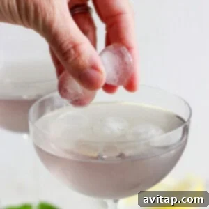 A hand pouring the mixed purple cocktail into a stemmed glass, leaving space for fresh ice and garnishes.