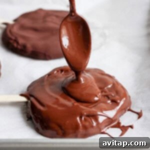 Delicious Homemade Dilly Bars 10 A spoon drizzles melted chocolate onto a round treat on a parchment-lined tray with a popsicle stick.